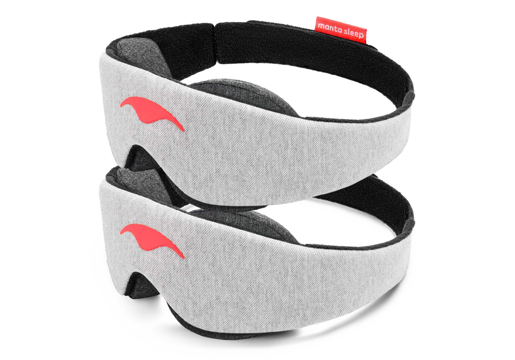 Two light gray heather fabric Manta Sleep Masks are stacked vertically against a clean white background. Both masks feature the signature smooth, coral-colored Manta Sleep logo on the center front and show a glimpse of the plush, dark gray contoured eyecups underneath the black-lined headband.