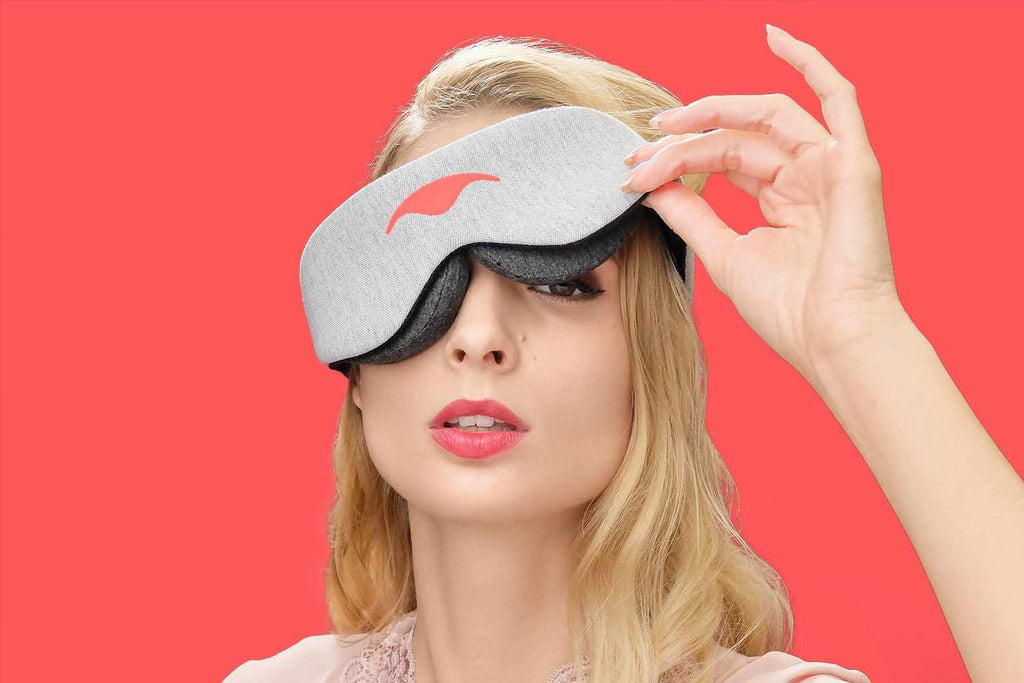 Close-up of a person lifting one side of a heather grey Manta Sleep Mask Classic against a solid vibrant red background.