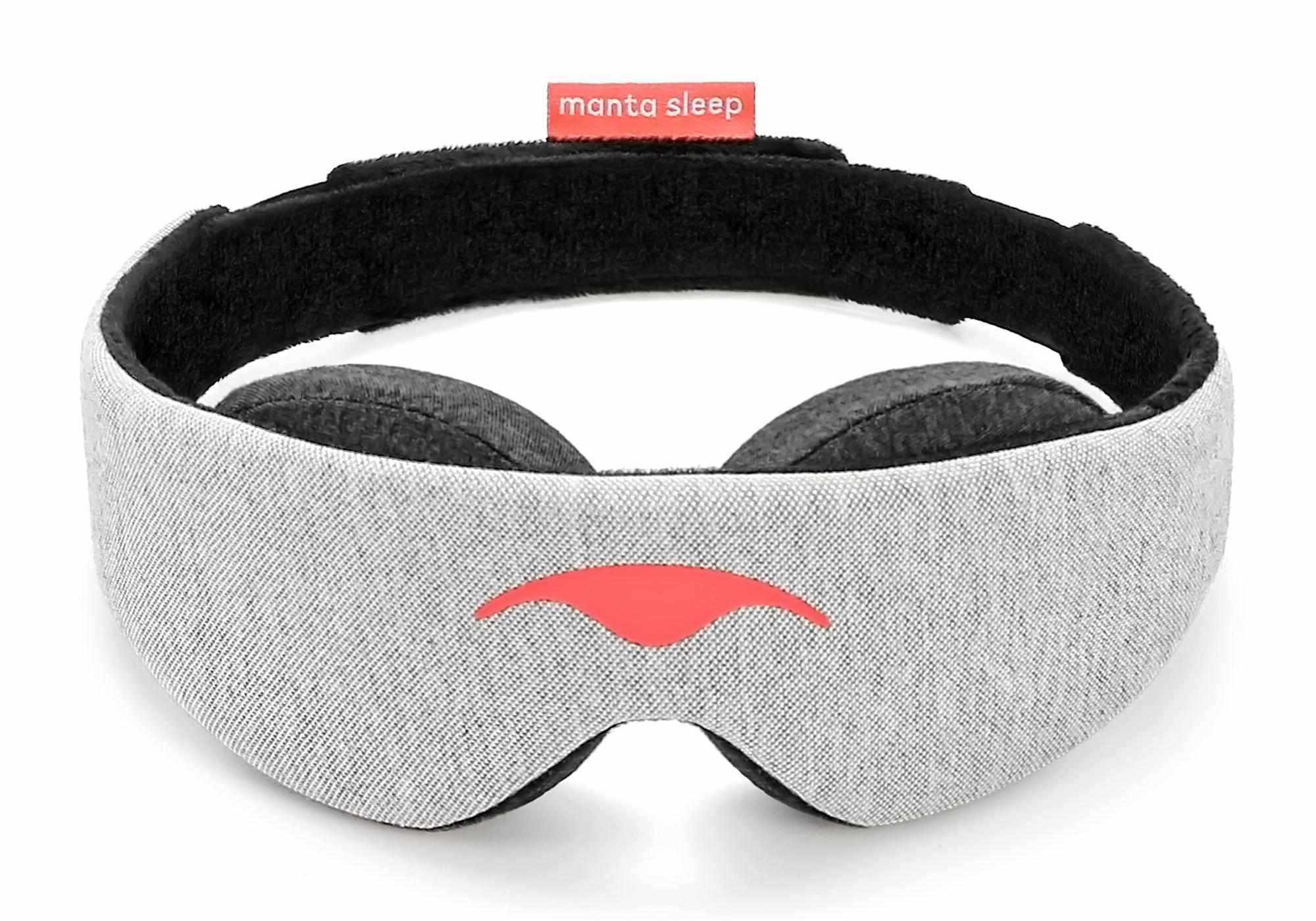 Front view of the Manta Sleep Mask in a circular, freestanding position against a white background. The main body is made of a light gray heathered knit fabric with a smooth, coral-red Manta Sleep logo printed in the center, contrasted by a black interior lining and a small coral brand tag at the back.