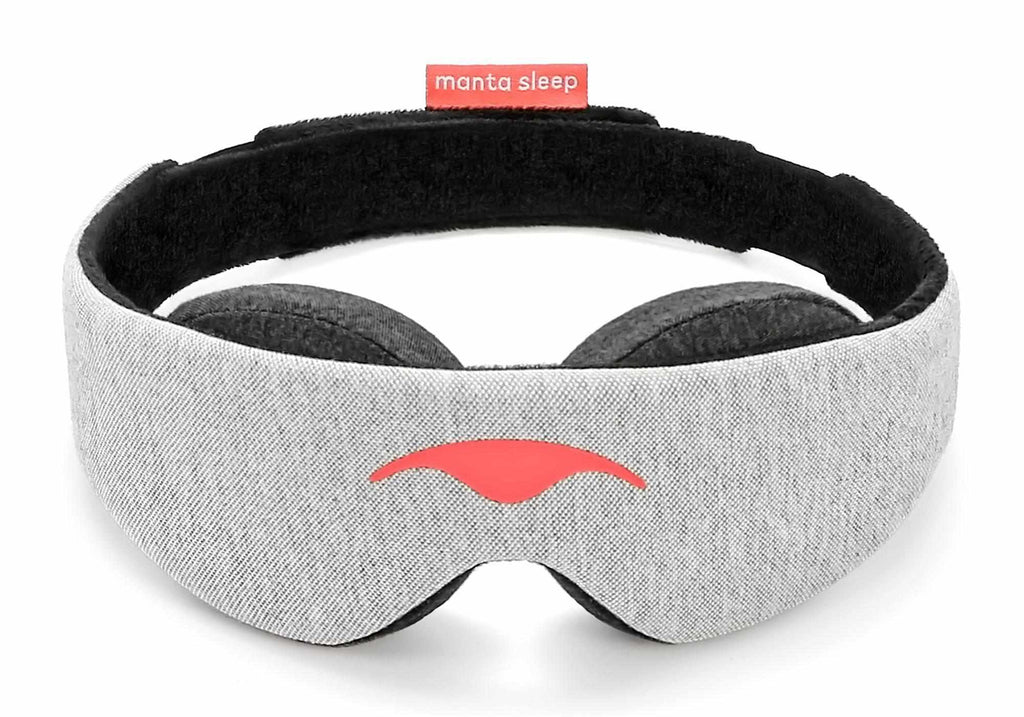 Front view of the Manta Sleep Mask in a circular, freestanding position against a white background. The main body is made of a light gray heathered knit fabric with a smooth, coral-red Manta Sleep logo printed in the center, contrasted by a black interior lining and a small coral brand tag at the back.