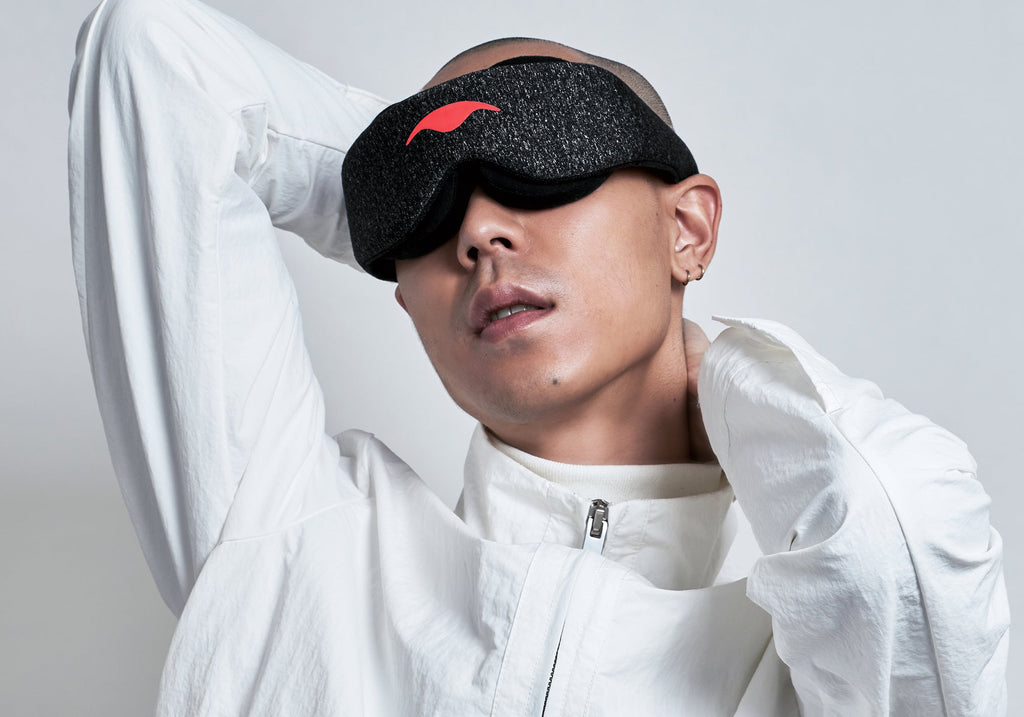 A studio portrait of a person with a shaved head wearing a dark gray heathered Carbon Black Manta Sleep Mask over their eyes. They are wearing a zippered white jacket and have their hands positioned behind their head and neck against a light gray backdrop.