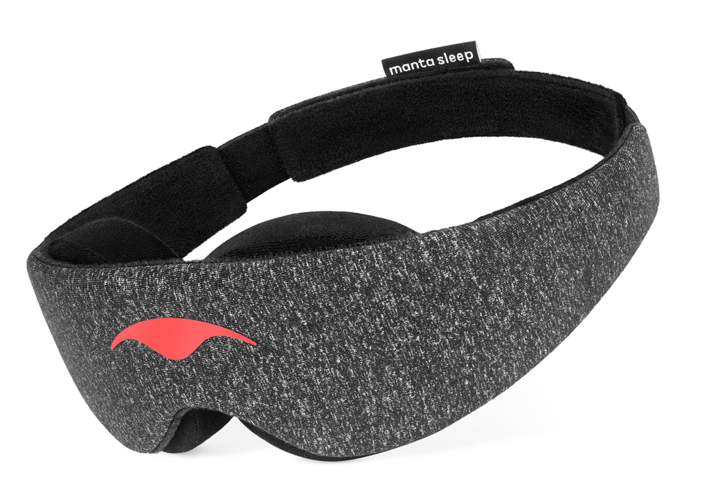 An angled product shot of the Carbon Black Manta Sleep Mask floating against a pure white background. The mask is constructed from dark gray speckled heather fabric with a smooth coral-red logo on the front, thick black contoured eye padding underneath, and a black brand tag on the rear strap.