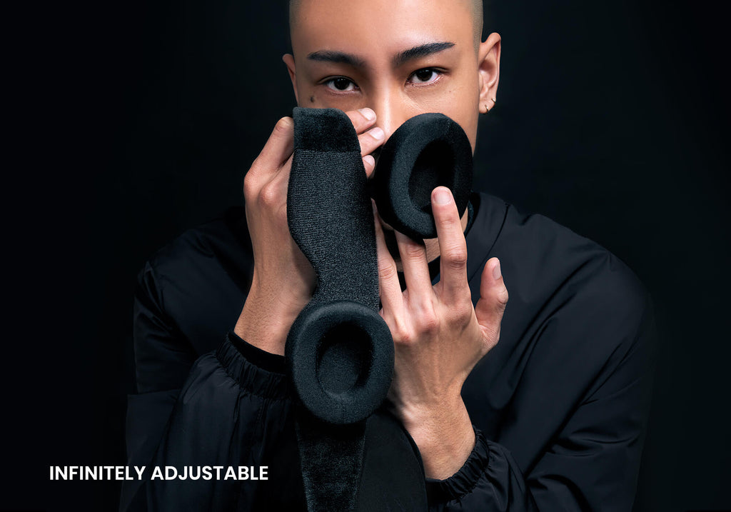 A close-up portrait of a person holding the detached black eyecup components of the sleep mask up to their face against a solid black background. The white text "Infinitely adjustable" is displayed in the lower left corner.