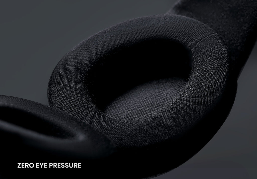 A macro, high-angle view focusing on the interior of a black sleep mask. It features a thick, contoured, oval-shaped eyecup covered in a smooth black knit material, with the white text "Zero eye pressure" located in the bottom left corner.