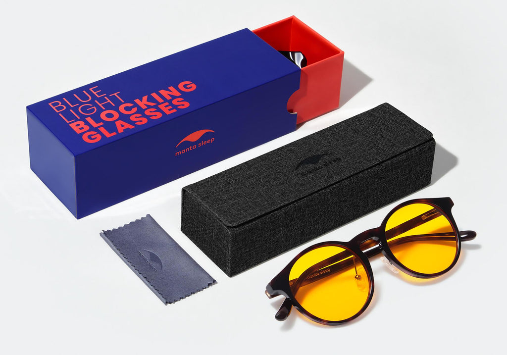 Manta blue light blocking glasses with yellow lenses next to a black hardshell case, a blue cleaning cloth, and a blue and red slide-out box.