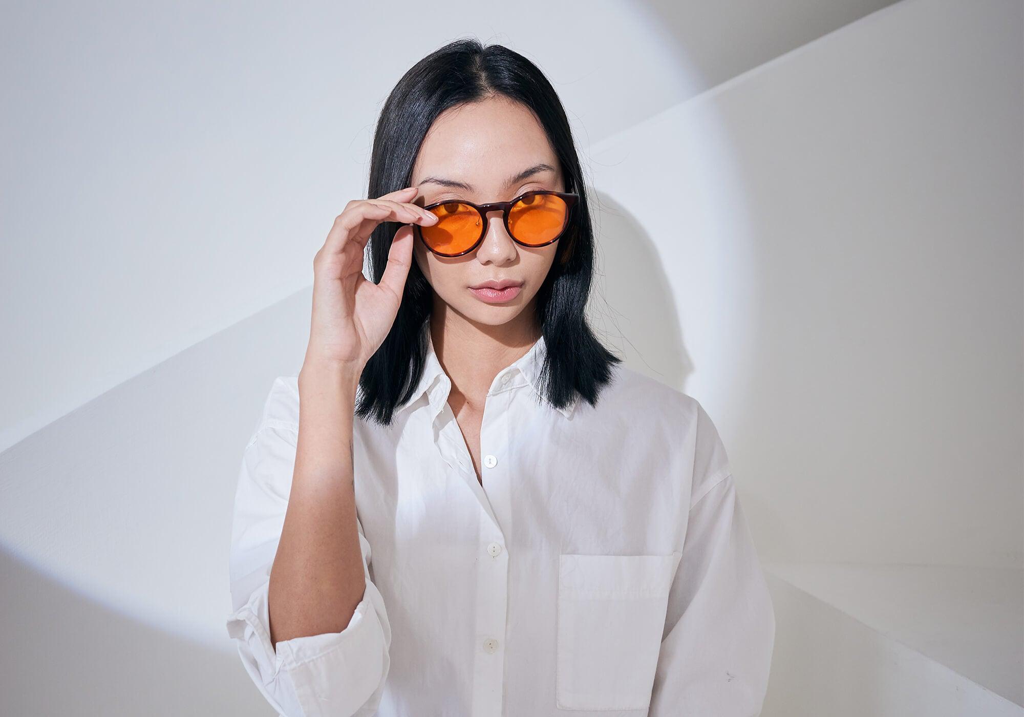 Frontal shot of a woman with dark hair in a white button-down shirt, adjusting her round tortoiseshell Manta blue light filtering glasses.