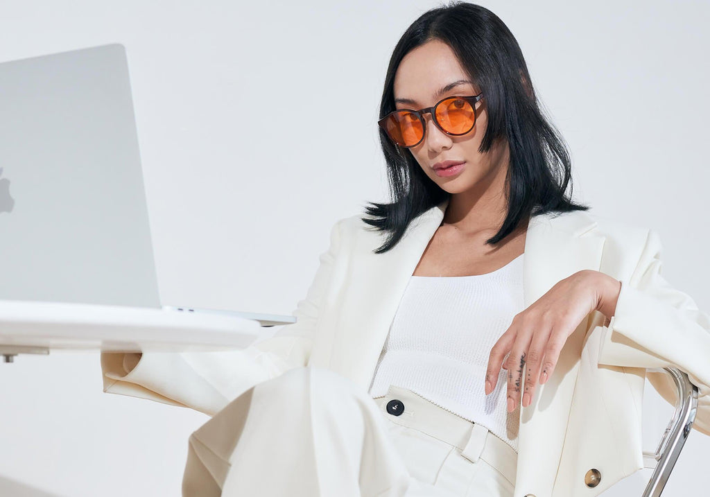 A woman with dark hair sits wearing a white blazer, white trousers, and orange-lensed blue light blocking glasses while looking down at her laptop.