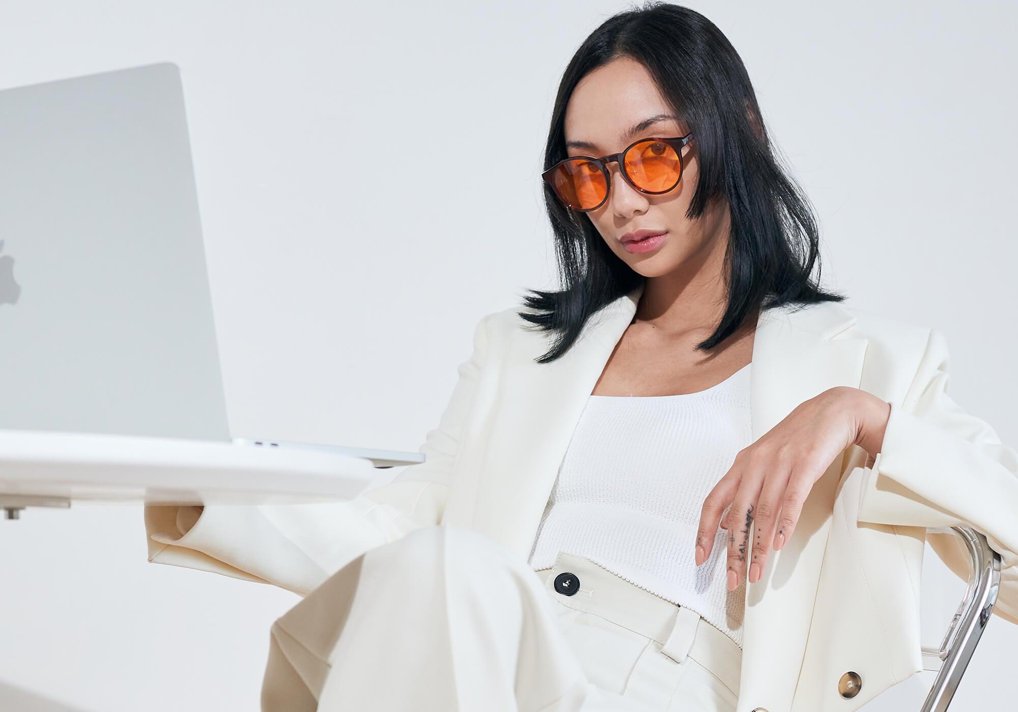 A woman with dark hair sits wearing a white blazer, white trousers, and orange-lensed blue light blocking glasses while looking down at her laptop.
