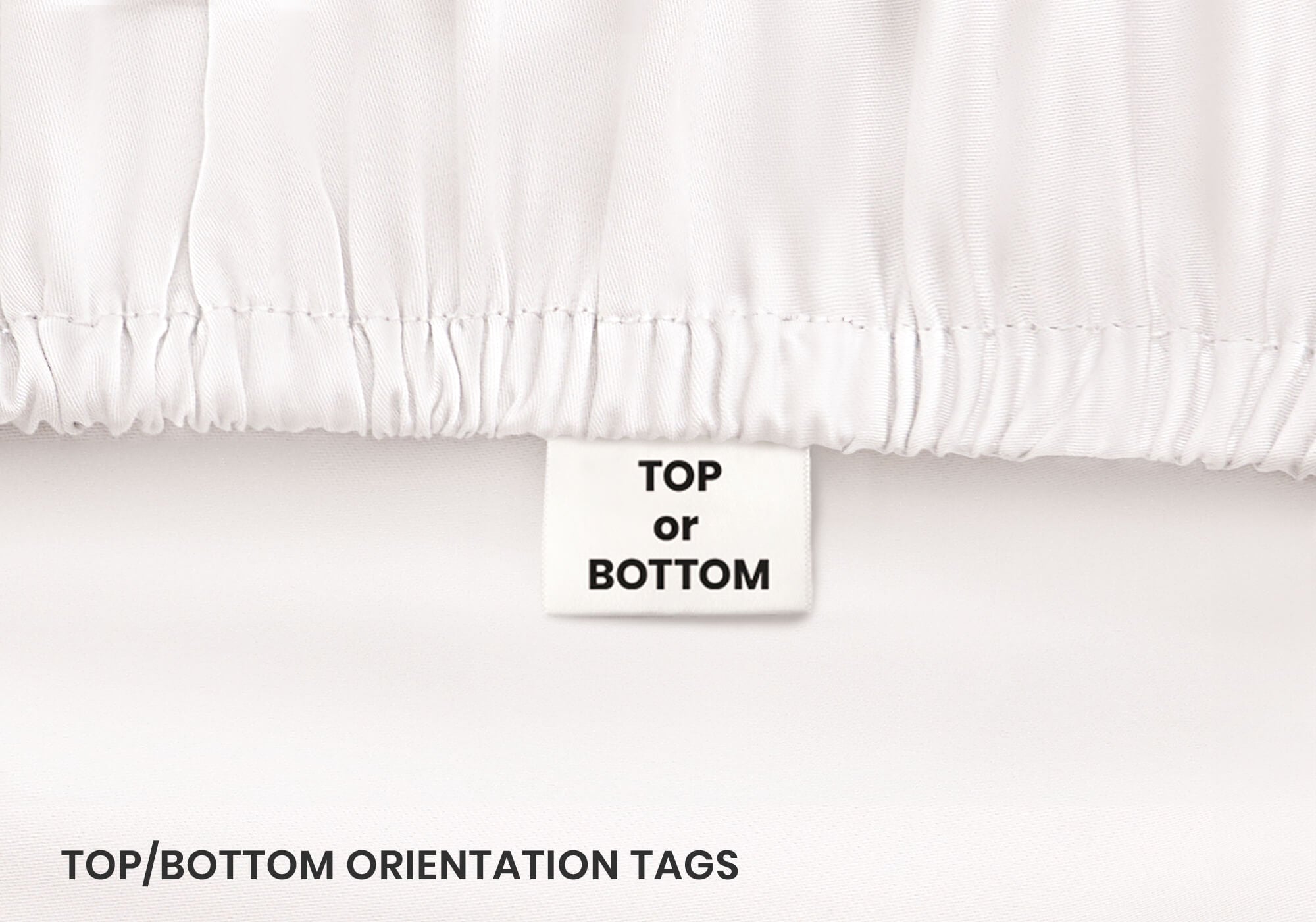 Close-up of a white fabric tag reading "Top or bottom" stitched onto a gathered white elastic sheet hem. Text overlay: "Top or bottom orientation tags".