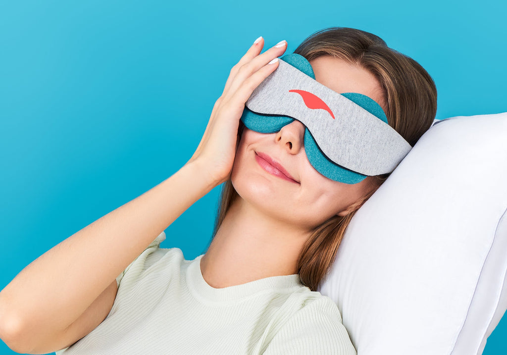 A woman resting her head on a white pillow while wearing a grey Manta COOL Sleep Mask fitted with blue-teal eye cups.
