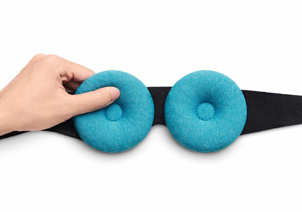 A hand adjusting a round, blue-teal fabric eye cup attached to sleep mask strap, isolated on white.