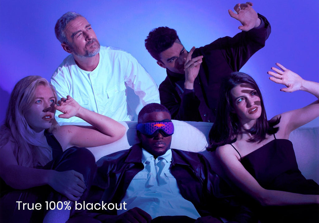 Five people on a couch under harsh light; one sits calmly wearing a purple Manta PRO Sleep Mask while the others shield their eyes from the glare, with text "True 100% blackout".