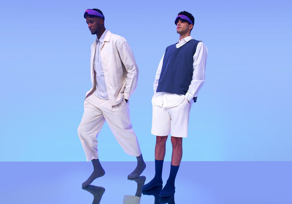 Two men modeling light-colored, casual outfits and wearing purple Manta Sleep Mask PROs on their foreheads against a light blue backdrop.