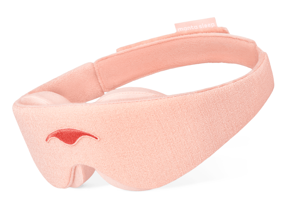 An angled product shot of the Sunrise Pink Manta Sleep Mask floating against a pure white background. The mask features a light pink soft-textured exterior, a darker pink embroidered logo on the front, thick contoured padding beneath, and a small brand tag on the upper back strap.