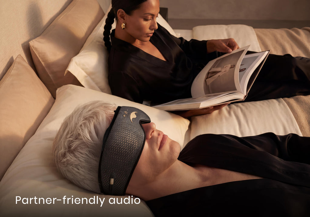 A man rests in bed wearing a grey audio mask next to a woman reading a magazine. Text overlay: "Partner-friendly audio".