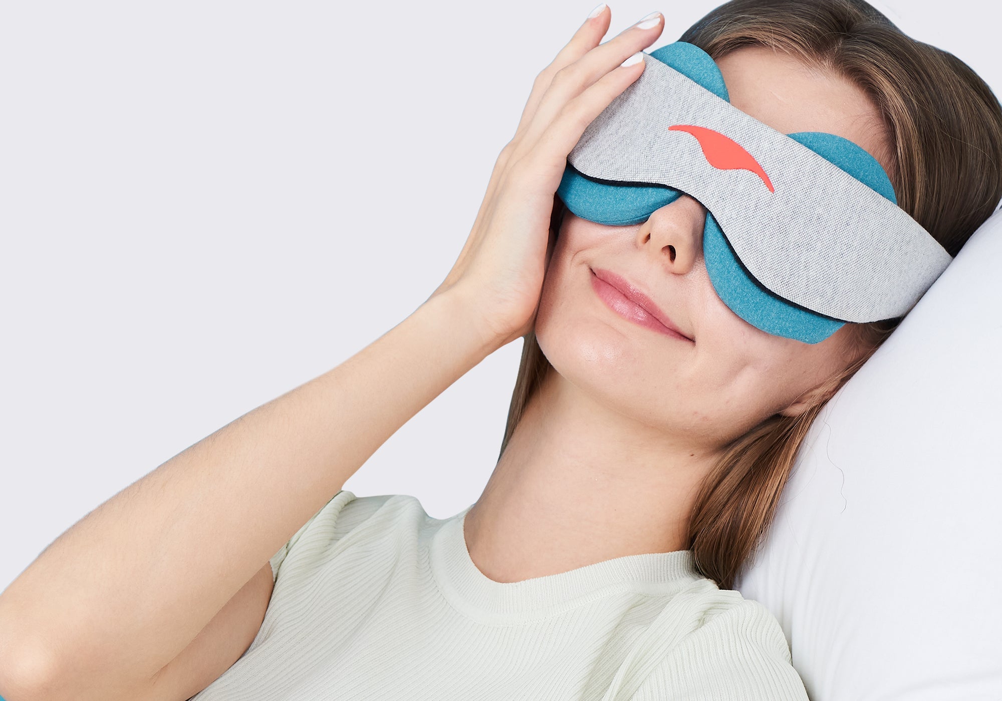 A woman resting her head on a white pillow while wearing a grey Manta COOL Sleep Mask fitted with blue-teal eye cups.