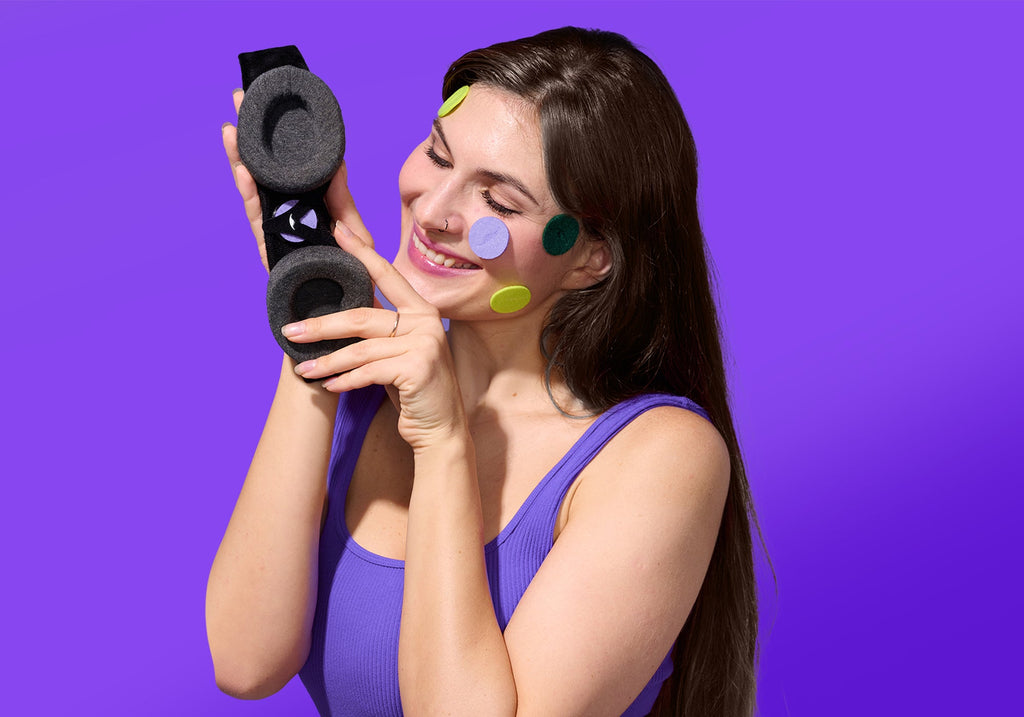 A smiling woman with colorful round aroma patches stuck on her face holds up the interior side of a Manta Sleep Mask, against a solid purple background.