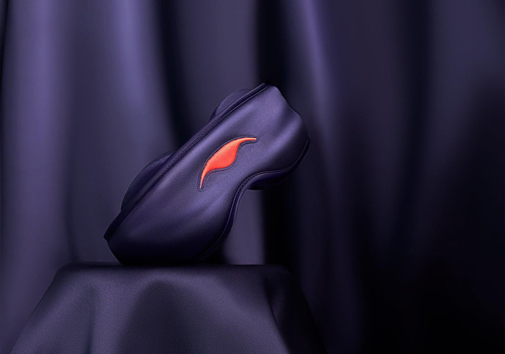 A purple silk sleep mask balancing elegantly on a pedestal draped in matching lustrous purple fabric.