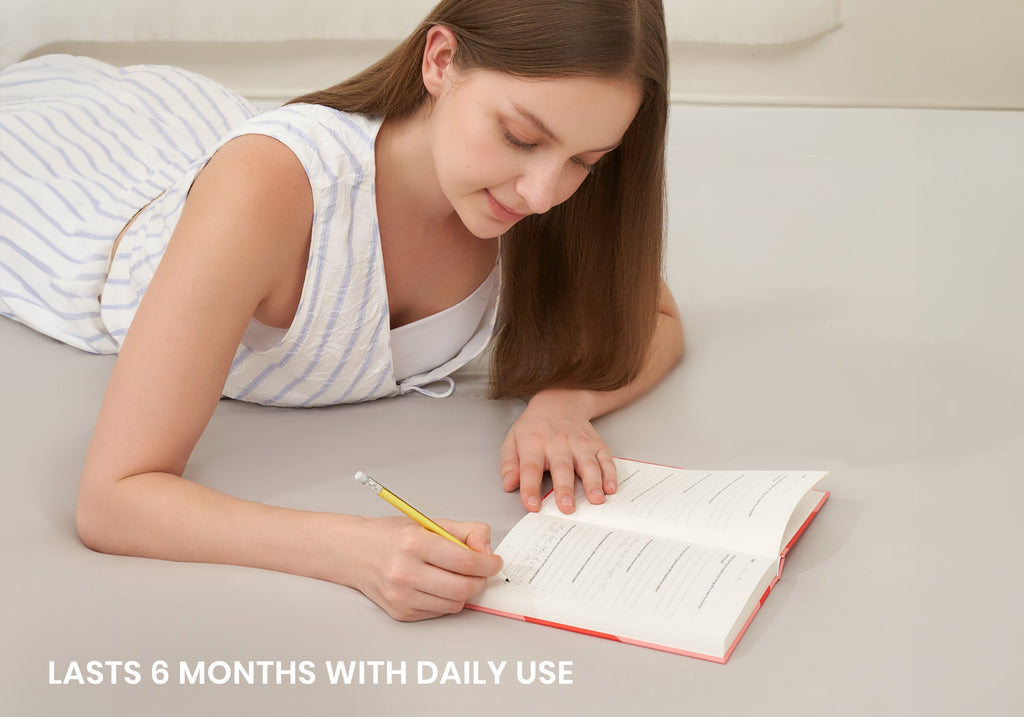 A woman lying on her stomach, writing with a yellow pencil inside an open Manta Sleep Journal. Overlay text reads "Lasts 6 months with daily use".