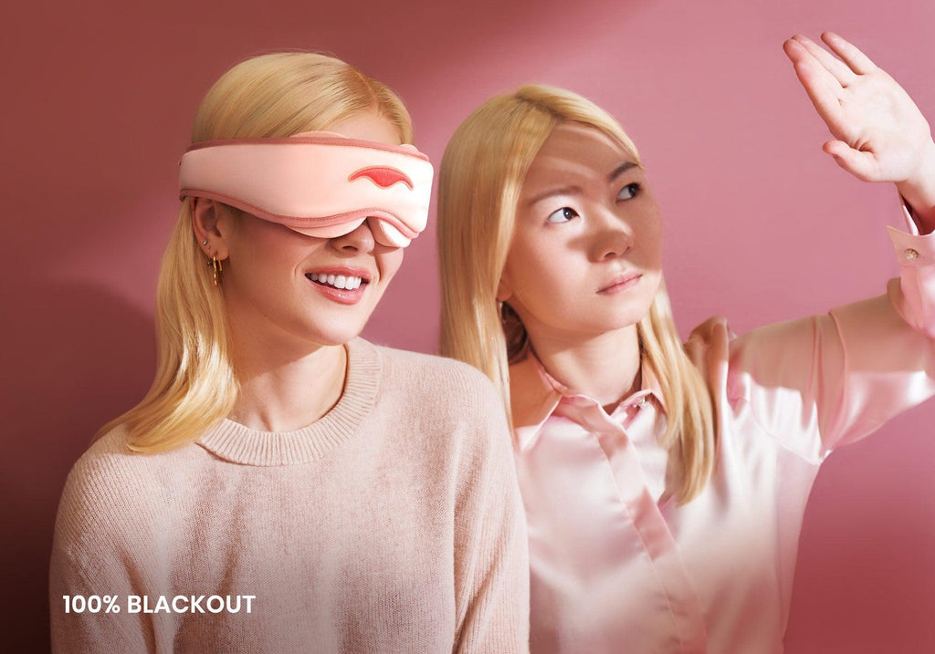 One person smiling in a pink sleep mask next to another shielding their eyes from bright light. Text overlay reads "100% blackout".