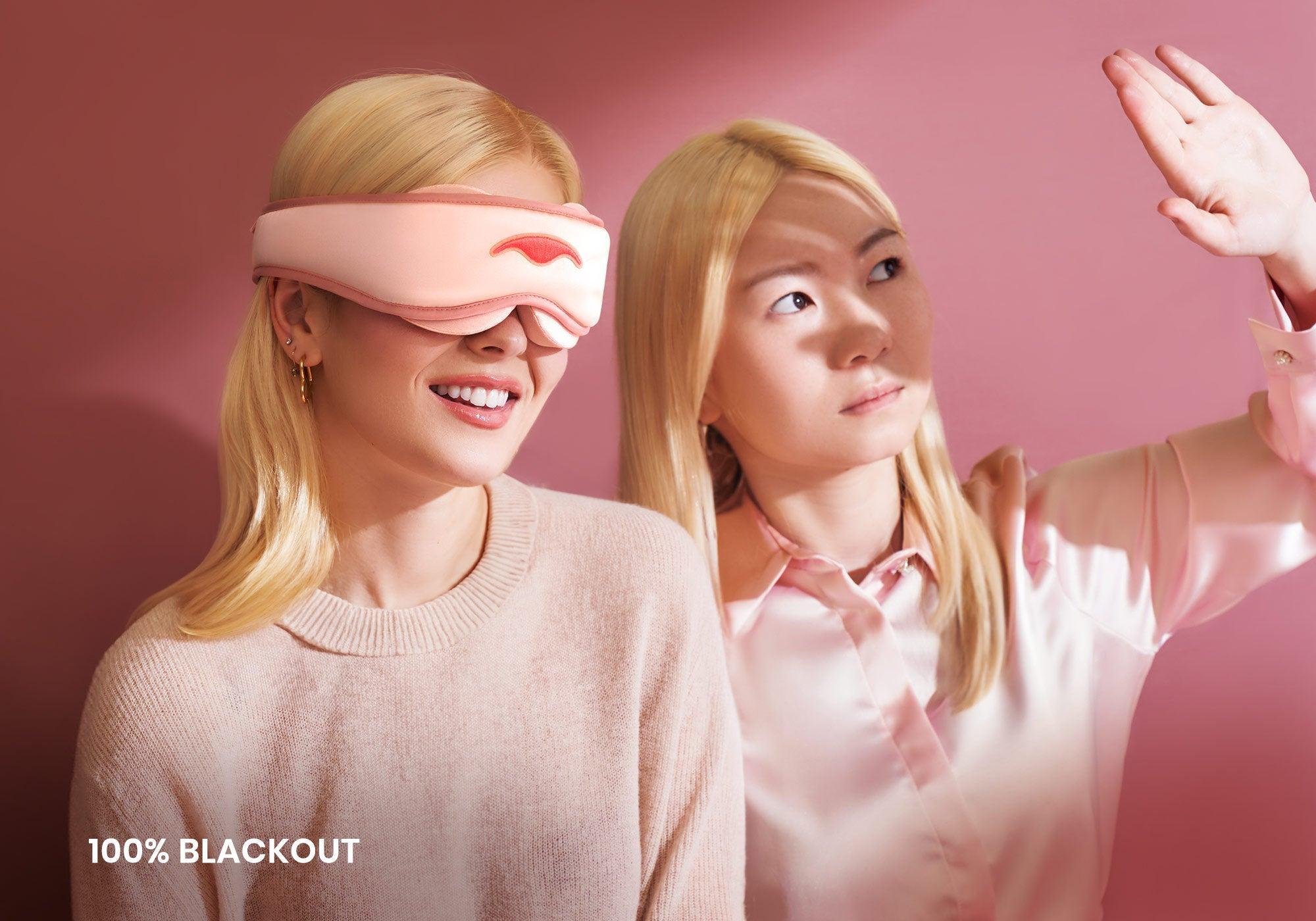 One person smiling in a pink sleep mask next to another shielding their eyes from bright light. Text overlay reads "100% blackout".