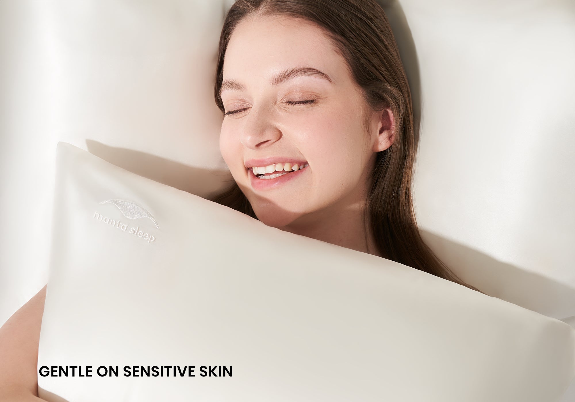 A smiling woman with closed eyes hugging a pearl white Manta Sleep silk pillowcase. Text overlay: "Gentle on sensitive skin".