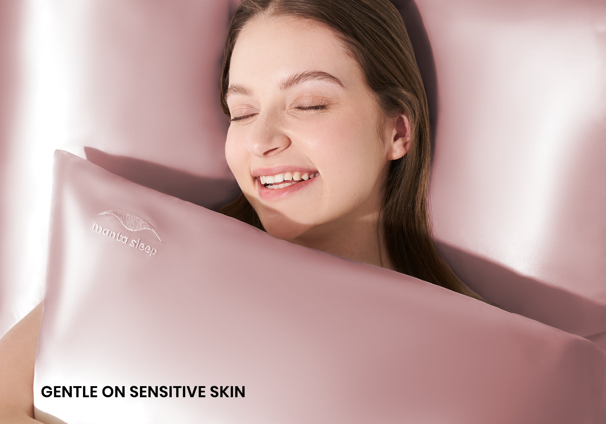 A smiling woman with closed eyes hugging a rose pink Manta Sleep silk pillowcase. Text overlay: "Gentle on sensitive skin".