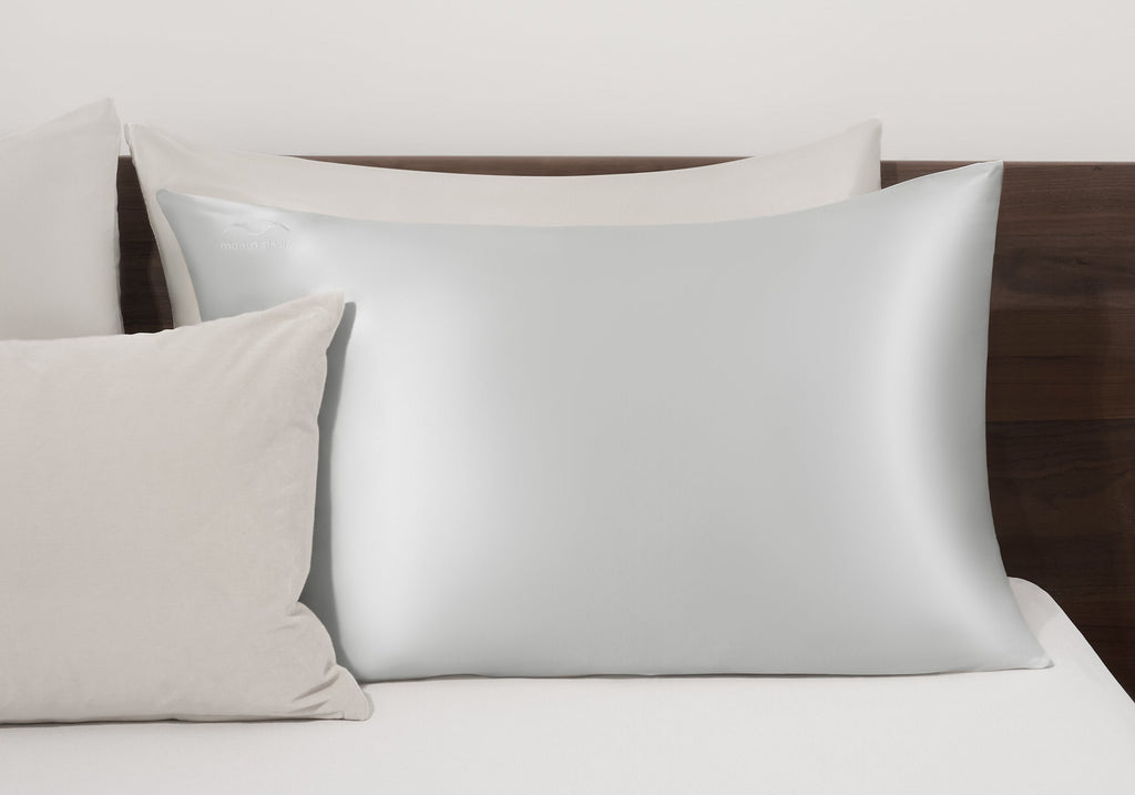 A crystal silver Manta Sleep silk pillowcase arranged on a bed with matching neutral-toned accent pillows against a wooden headboard.