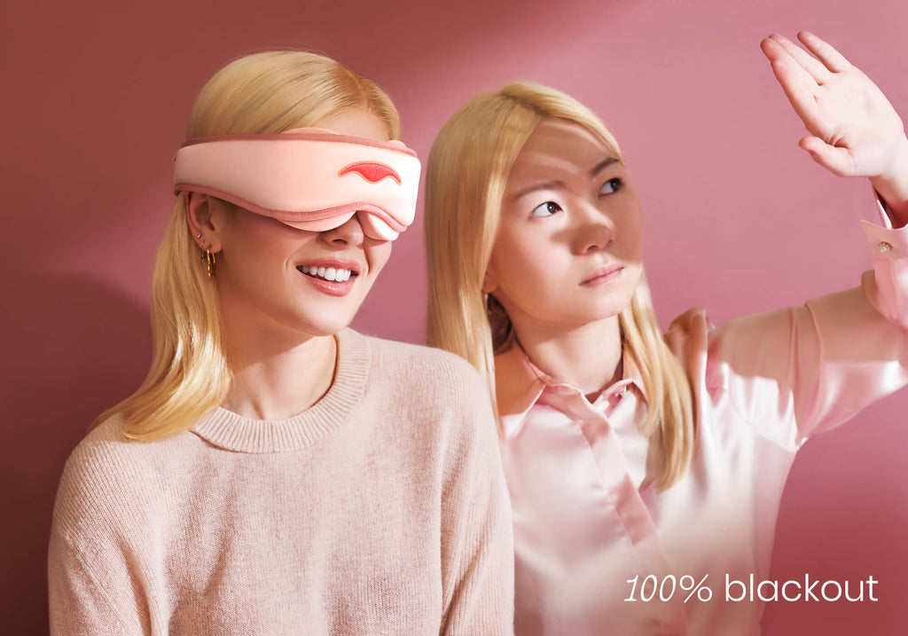One person smiling in a pink sleep mask next to another shielding their eyes from bright light. Text overlay reads "100% blackout".