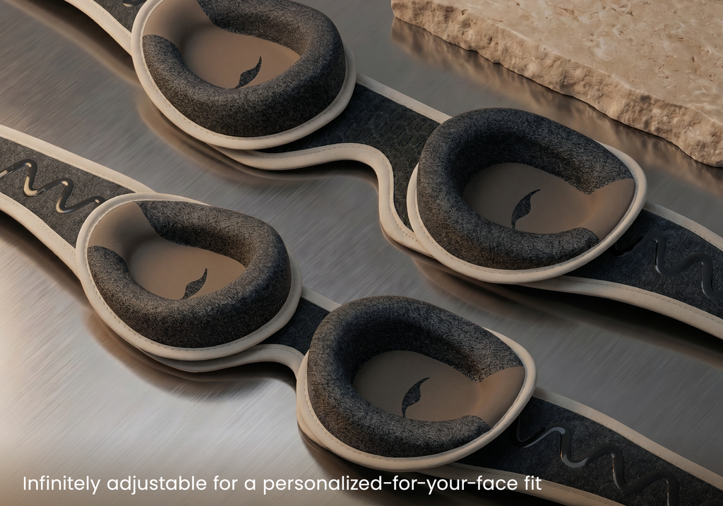 Close-up of a pair of Tranquil Taupe Manta PRO Sleep Mask, shown on a brushed metal surface with a stone slab in the background. Text at the bottom reads "Infinitely adjustable for a personalized-for-your-face fit".