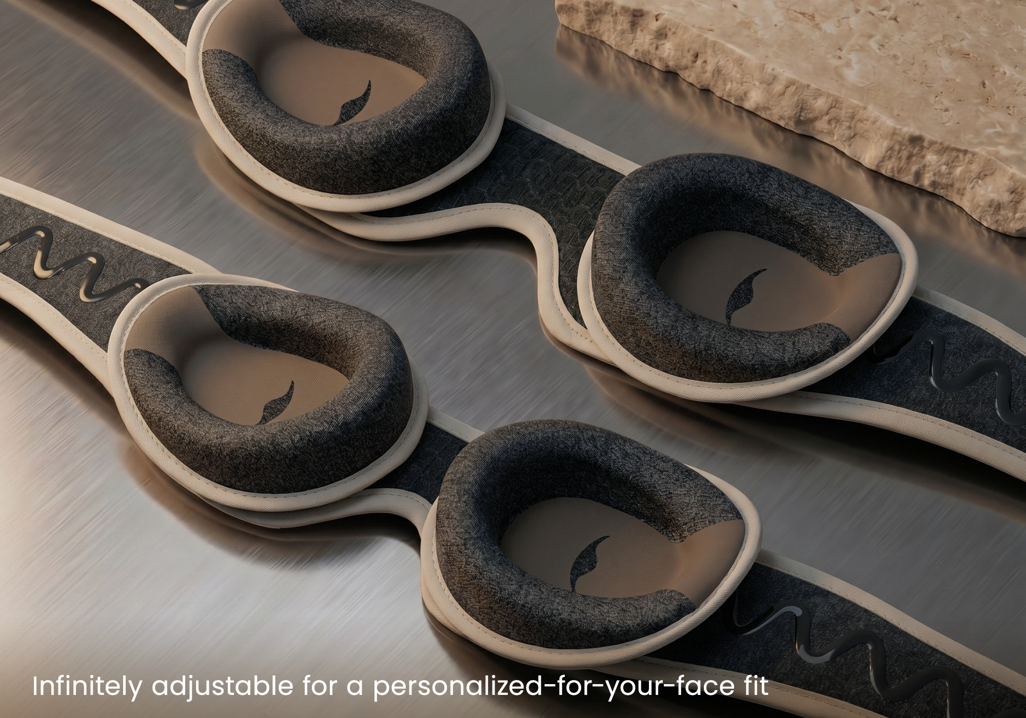 Close-up of a pair of Tranquil Taupe Manta PRO Sleep Mask, shown on a brushed metal surface with a stone slab in the background. Text at the bottom reads "Infinitely adjustable for a personalized-for-your-face fit".