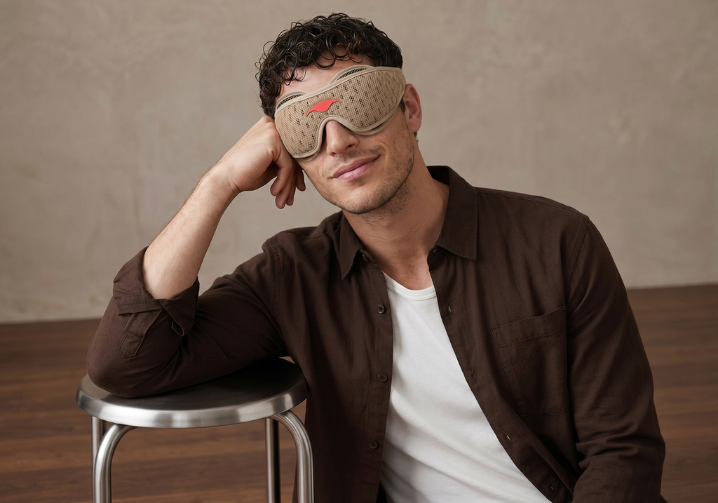 A man wearing a Tranquil Taupe Manta PRO Sleep Mask and a brown button-up shirt over a white t-shirt, leaning on a metal stool against a textured beige wall.