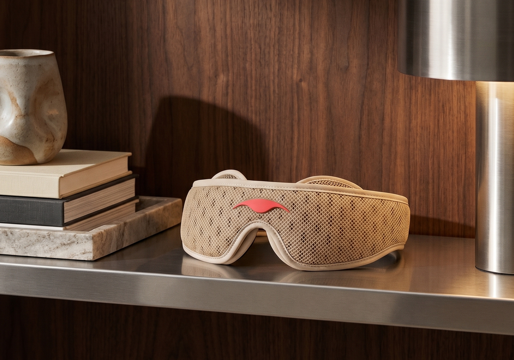 A Tranquil Taupe Manta PRO Sleep Mask, resting on a metallic shelf next to books and a lamp against a dark wood background.