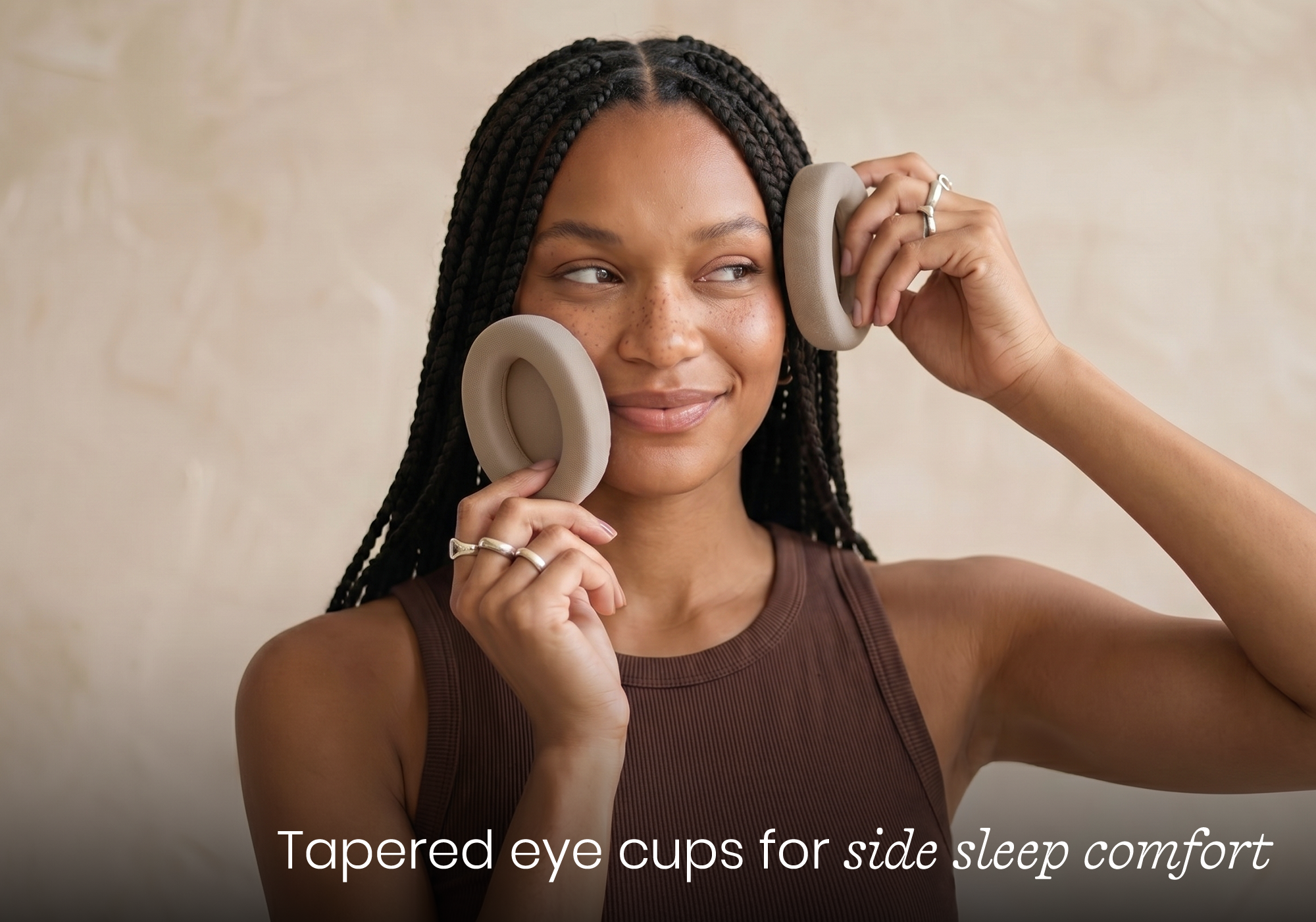 A woman with braided hair holds Tranquil Taupe tapered eye cups to her eyes, demonstrating their use for side sleep comfort.