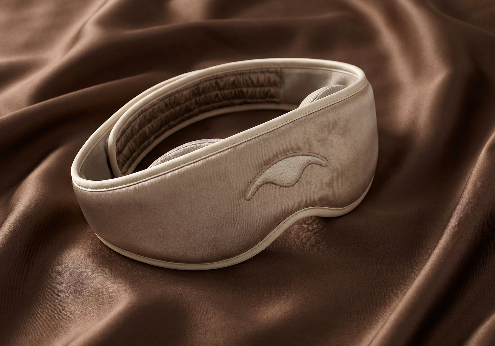 A Tranquil Taupe Manta SILK Sleep Mask, resting on a draped brown satin fabric background.