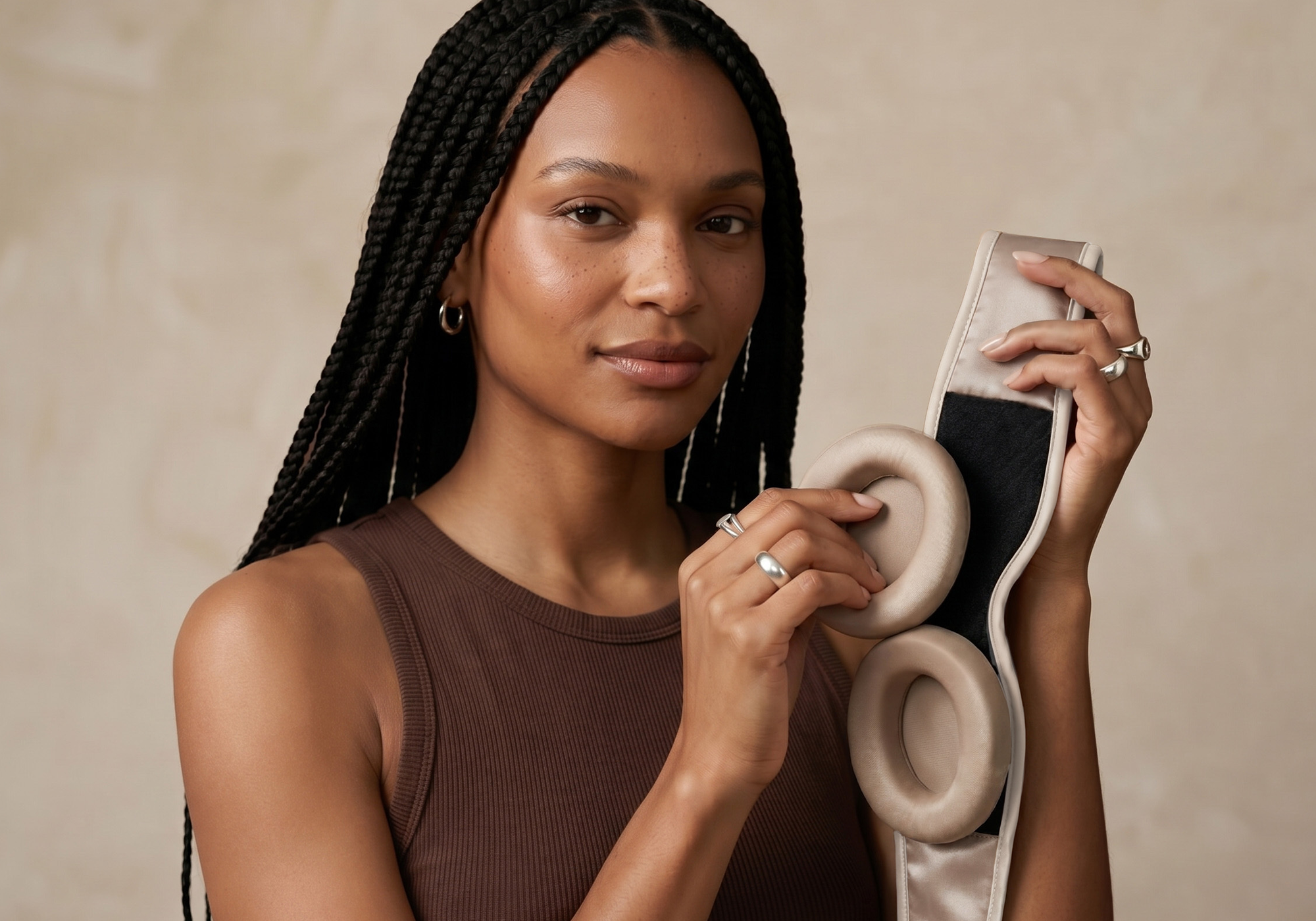 A woman with long black braids holds a eye cup and Tranquil Taupe Manta SILK Sleep Mask against a neutral beige background.
