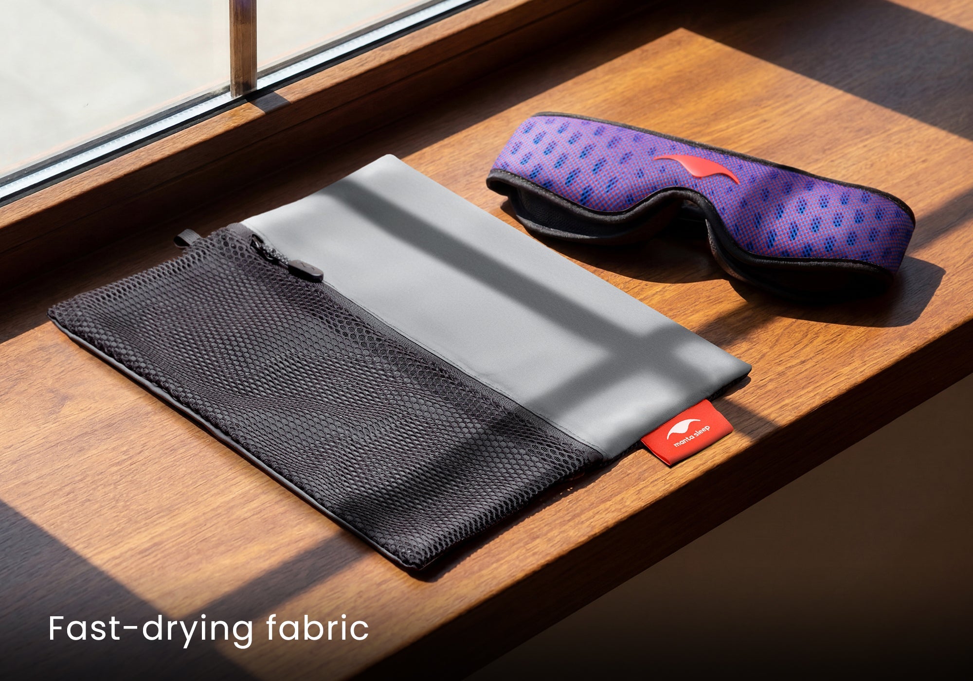 An unfolded grey and black mesh Manta Travel & Laundry Bag resting on a wooden window sill next to a blue and red Manta PRO Sleep Mask in the sunlight. Text overlay: "Fast-drying fabric".