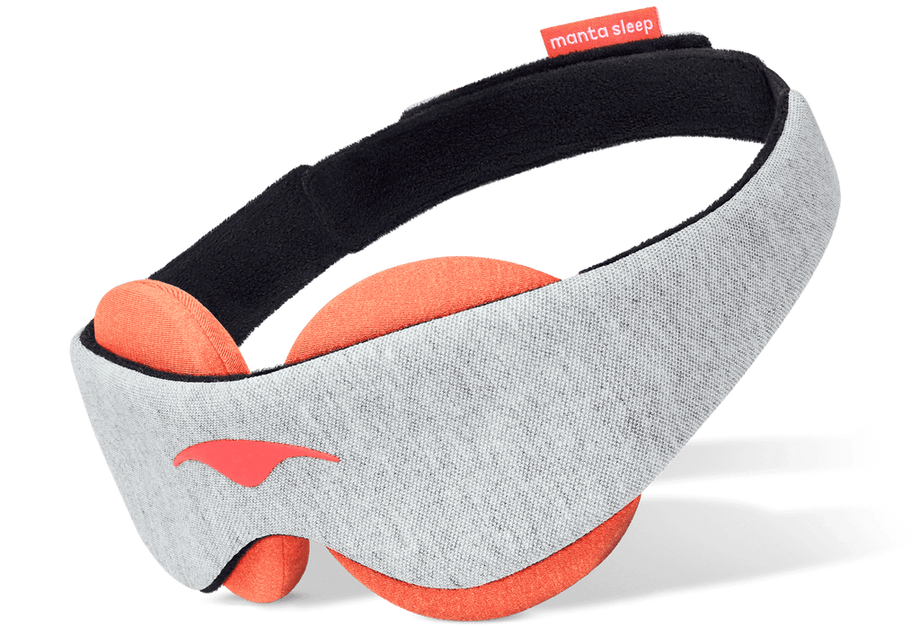An angled product shot of the Manta STEAM Sleep Mask floating against a crisp white background. The exterior body displays a soft light gray heathered fabric with a coral-pink Manta Sleep logo on the left profile, while the interior reveals thick circular terracotta-orange eye cups designed for heat therapy, attached to a black padded head strap.