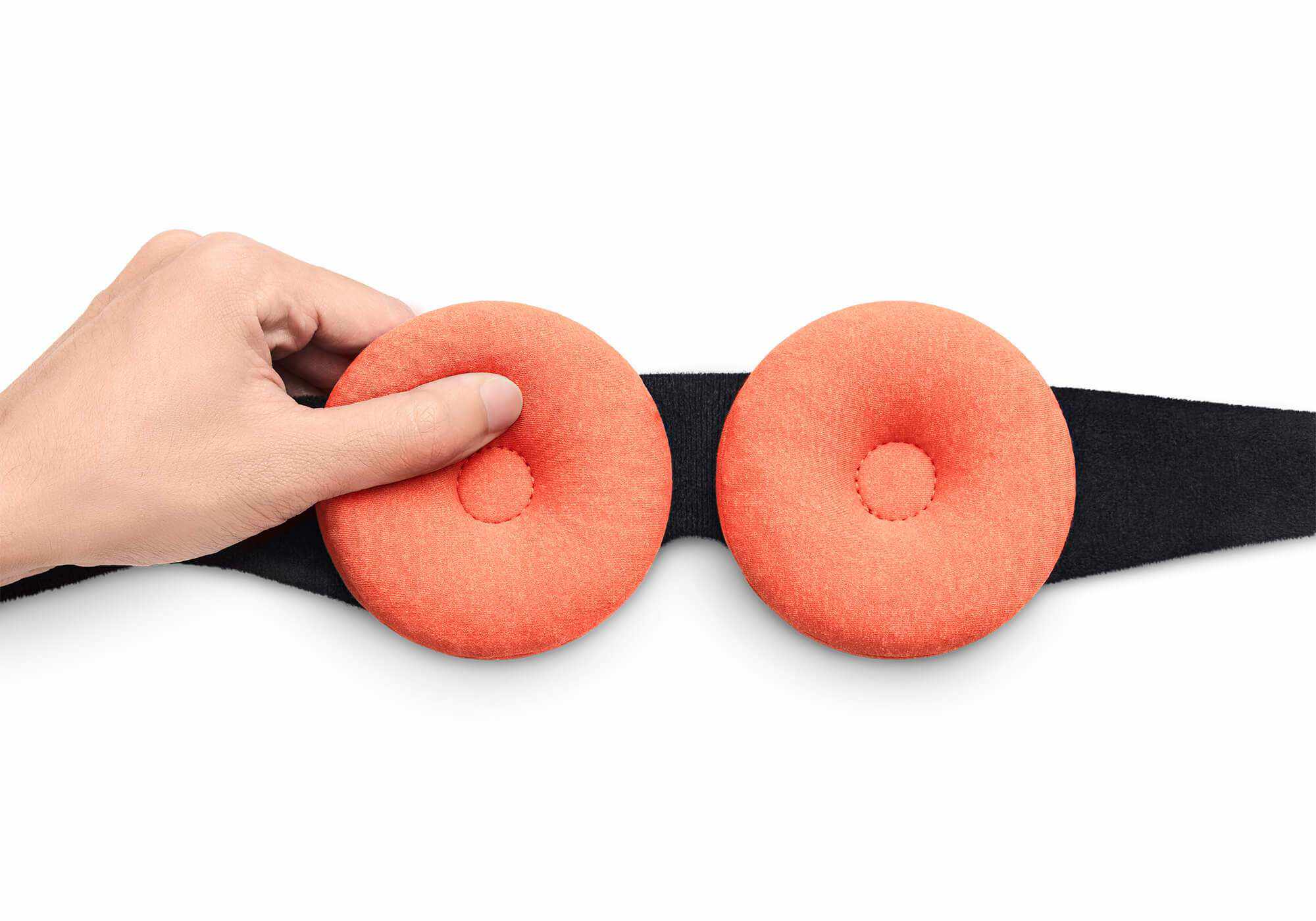 A hand attaching a round orange fabric eye cup onto a sleep mask strap, isolated on a white background.