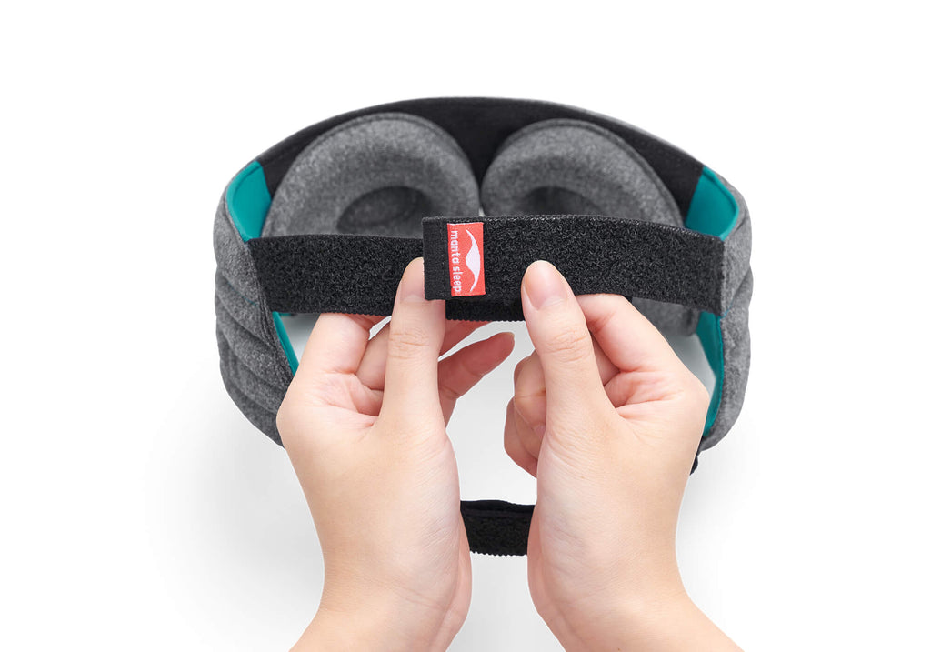 A close-up view of two hands adjusting the micro-hook velcro closure at the back of the Manta WEIGHTED Sleep Mask. The black elastic strap features a small red tag with the white "manta sleep" logo, while the heather-gray eye cups and bright teal side linings are visible in the background against a clean white surface.