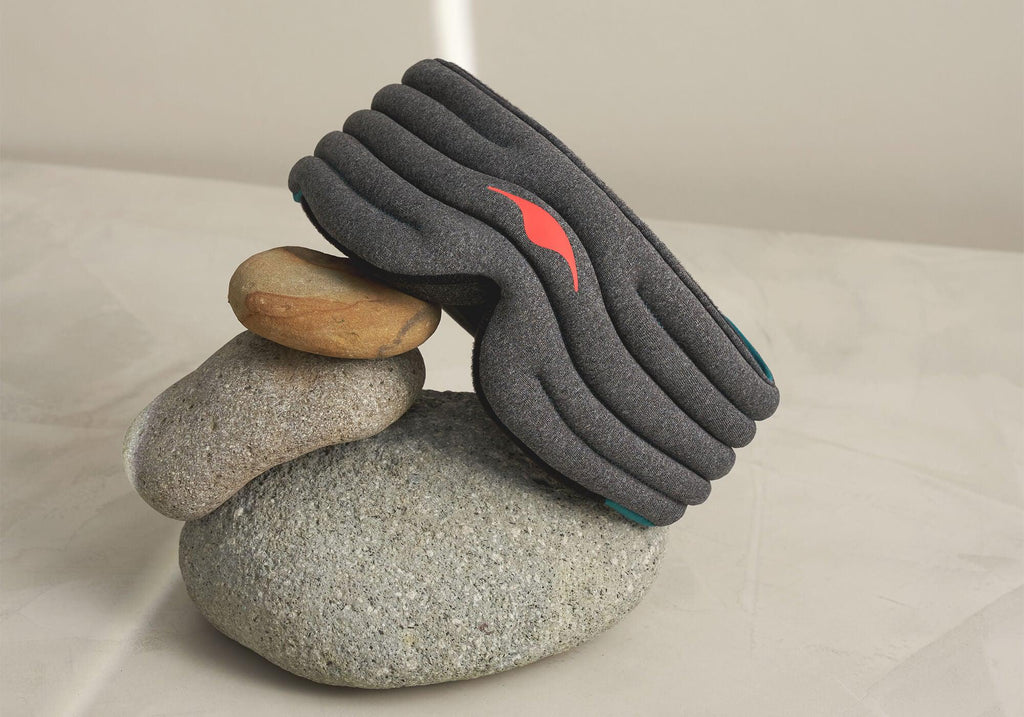 A creative product shot featuring the heather-gray Manta WEIGHTED Sleep Mask balanced gracefully atop a small stack of three smooth, natural river stones. The scene is illuminated by soft light on a neutral surface, showcasing the quilted rows of the mask and its red emblem.