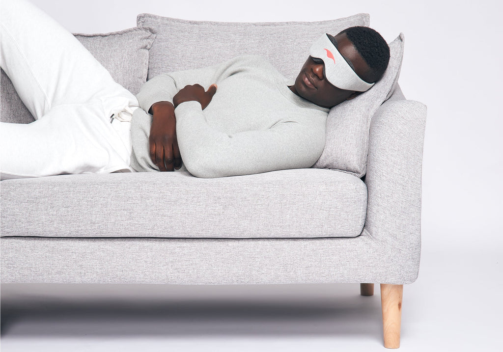 A person relaxing on a light grey fabric sofa while wearing a heather grey Manta Sleep Mask Classic.