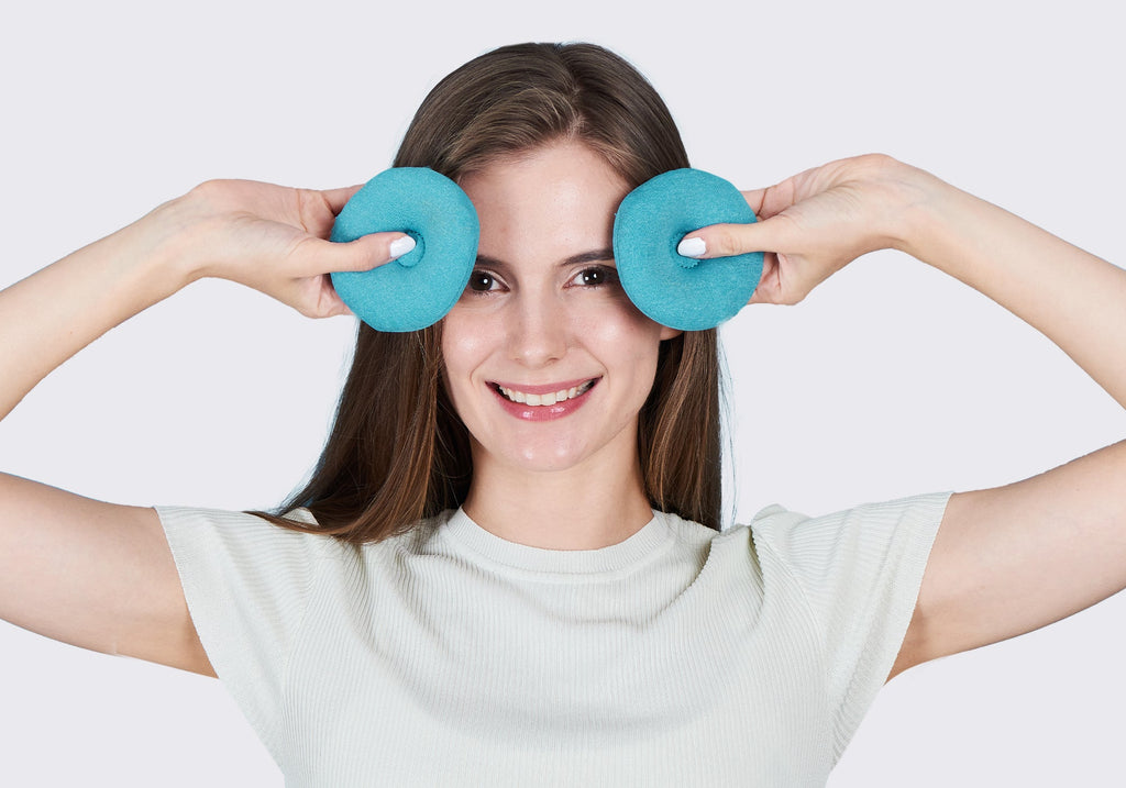 A smiling woman holding two round, blue-teal fabric eye cups over her eyes against a plain light gray background.
